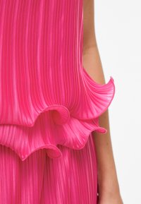 Φωτεινό ροζ πλισέ ύφασμα με βολάν σε στρωματοποιημένες άκρες, φορεμένο στον άνω βραχίονα και στον κορμό ενός ατόμου, με φόντο λευκό.
