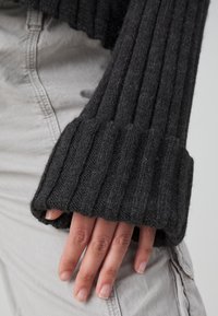 Main aux ongles naturels partiellement couverte par une longue manche en tricot côtelé gris foncé, portée sur un pantalon gris clair.