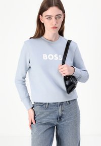 Gaiši zila džemperis ar baltu "BOSS" logotipu, apaļa kakla izgriezums, garas piedurknes, kopā ar augstas jostasvietas zilām džinsām un melnu somiņu.