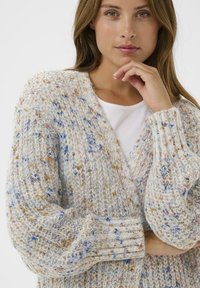 Junge Frau mit braunem Haar, die einen mehrfarbigen, gepunkteten Strickcardigan über einem weißen Oberteil trägt und ihr Kinn auf ihre Hand stützt.