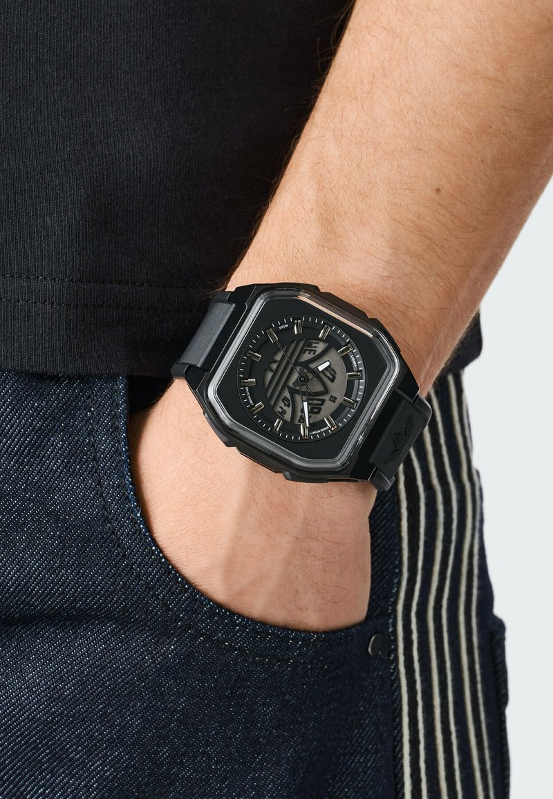 Black digital and analog wristwatch on left wrist with hand in dark denim jeans pocket and black shirt sleeve.