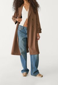 Abrigo marrón de doble botonadura sobre una camiseta blanca de canalé, combinado con jeans de pierna ancha en azul claro y zapatillas beige con una superficie texturizada.