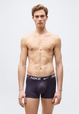 Νέος άντρας με σγουρά μαλλιά που φοράει μαύρα εσώρουχα Nike boxer, στέκεται μπροστά σε απλό ανοιχτό γκρι φόντο, κοιτάζοντας προς τα εμπρός.