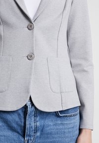 Blazer gris claro con un diseño ajustado, dos bolsillos frontales y dos botones redondos. Hecho de una tela suave, combinado con jeans de mezclilla azules.