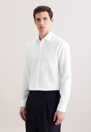 Homme aux cheveux bruns courts portant une chemise blanche à manches longues boutonnée et un pantalon foncé, debout devant un fond clair uni.