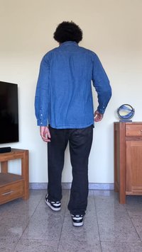 Džínová košeľa v modrej farbe, s dlhými rukávmi, gombíkovými manžetami a zakriveným spodným okrajom. Nosená s tmavými nohavicami a čiernymi teniskami. Jednoduché pozadie.