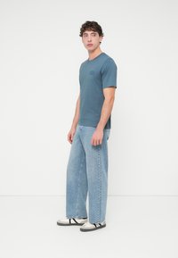 Μπλε μπλουζάκι με ένα μικρό μαύρο λογότυπο, συνδυασμένο με ανοιχτό μπλε τζιν παντελόνι φαρδιάς γραμμής και γκρι αθλητικά παπούτσια με μαύρα λουράκια. Το μοντέλο στέκεται μπροστά σε λευκό φόντο.