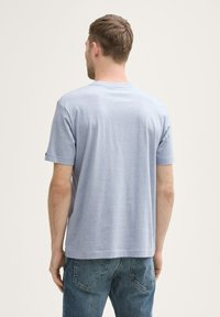Bărbat cu părul scurt, maro, purtând un tricou cu mânecă scurtă de culoare albastră deschisă și blugi, stând cu spatele, pe un fundal simplu.