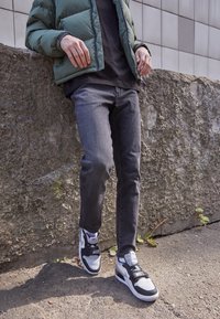 Vaqueros de mezclilla negros y una chaqueta acolchada verde, combinados con zapatillas blancas y negras con una correa, frente a una pared de piedra texturizada.