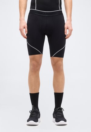 Uomo che indossa pantaloncini neri compressivi con piping riflettente, calzini neri e scarpe da ginnastica nere su uno sfondo semplice.