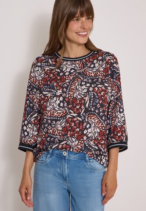 Femme souriante portant un chemisier multicolore à motifs floraux et abstraits avec des manches trois quarts et un jean bleu clair.