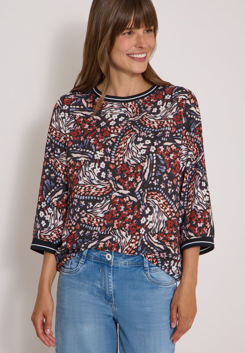 Femme souriante portant un chemisier multicolore à motifs floraux et abstraits avec des manches trois quarts et un jean bleu clair.