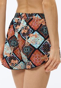 Γυναίκα που φοράει σορτς με μοτίβα γεωμετρικά και λουλουδάτα σε κόκκινο, μπλε, μαύρο και μπεζ, με θέα από πίσω και πλάγια.