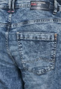 Denim jeans met een lichte blue-wash, voorzien van een achterzak met een gestikte detail en een metalen accent. Het label zegt "ONTDEKKEN VAN PURE VREUGDE."