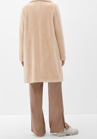 Kvinna som bär en beige mjuk pälsjacka, ljusbruna vida byxor med slitsar vid sidorna och vita klumpiga loafers står mot en vit bakgrund.