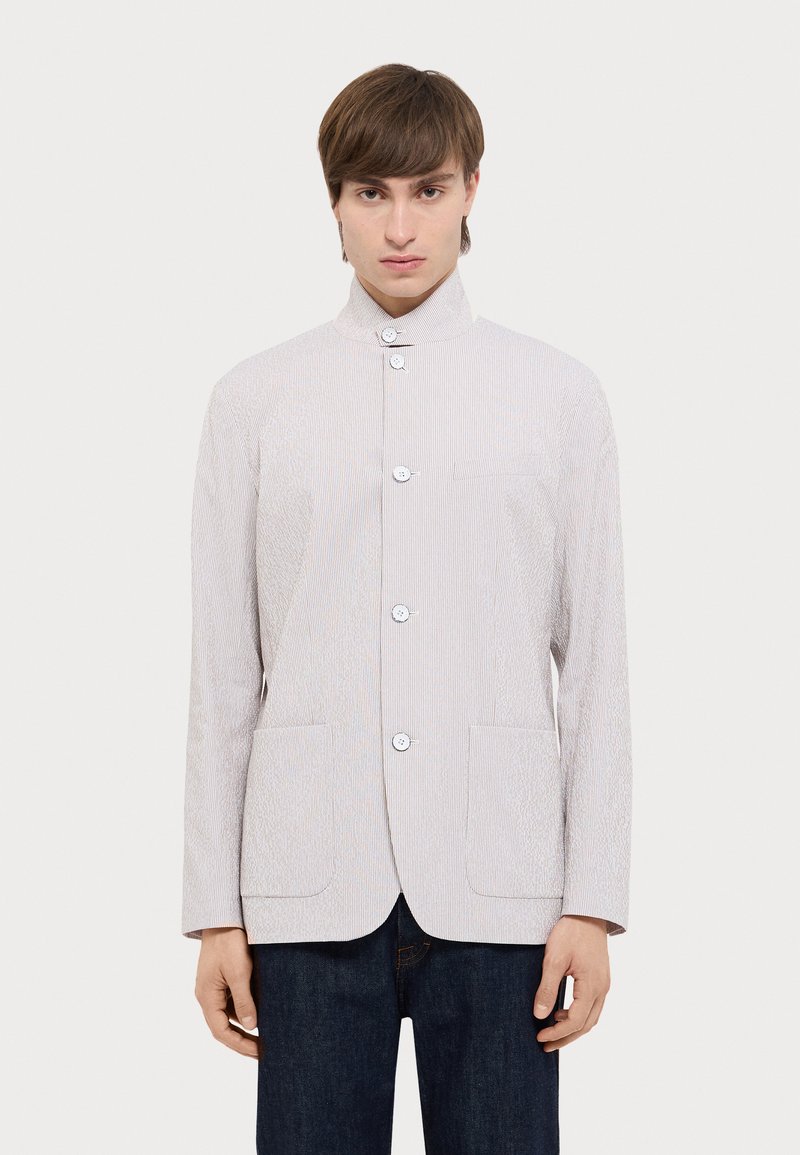 Helle graue Jacke mit hochgeschlossenem Kragen, horizontalen Streifen und vier Knöpfen. Verfügt über zwei Fronttaschen und einen insgesamt strukturierten Stoff.