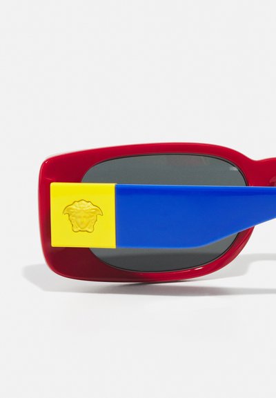 Red sunglasses with dark lenses, featuring blue and yellow temple accents. Yellow accent has a textured logo. Smooth plastic material.