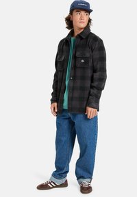 Camisa de franela a cuadros en negro y gris con cierre de botones y dos bolsillos en el pecho, llevada sobre una camisa verde, combinada con jeans azules holgados y zapatos marrones.