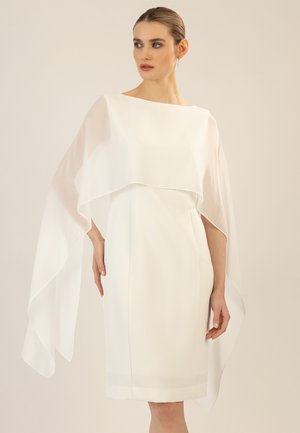 Frau trägt ein weißes knielanges Kleid mit einem durchsichtigen Cape-Überwurf, hat die Haare zurückgebunden und trägt hängende Ohrringe.