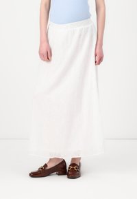 Jupe longue blanche avec des détails en broderie anglaise, taille élastique, associée à des mocassins en cuir verni brun avec un accent doré décoratif.