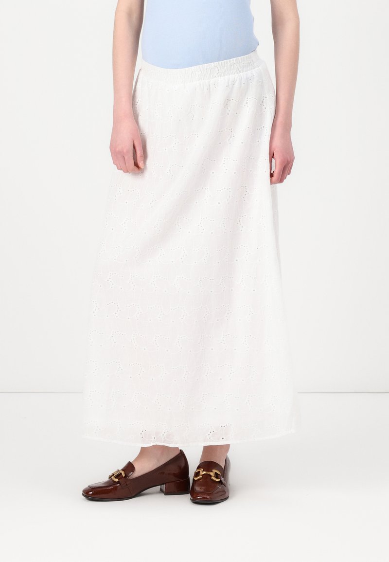 Jupe longue blanche avec des détails en broderie anglaise, taille élastique, associée à des mocassins en cuir verni brun avec un accent doré décoratif.