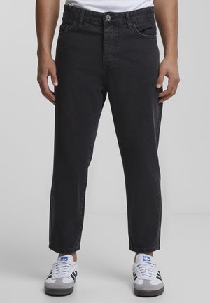 Personne portant un jean noir et des baskets blanches Adidas avec un logo bleu, debout devant un fond gris uni.