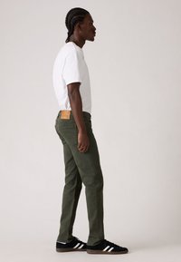 Pantaloni verde oliva con taglio dritto, tessuto testurizzato e una toppa in pelle sul retro. Indossati con una t-shirt bianca e sneaker nere.