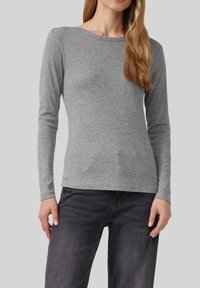 Woman wearing a fitted long-sleeve grey shirt and dark grey jeans, standing against a plain light grey background.