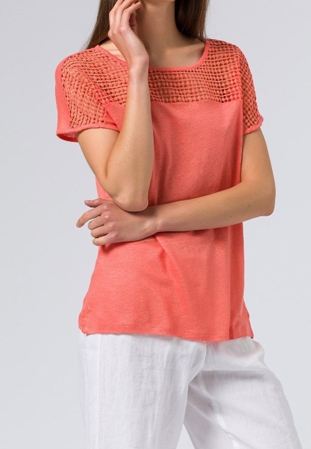 Coral short-sleeve top with a textured, open-knit pattern on the shoulders, featuring a relaxed fit and smooth fabric. Paired with white trousers.