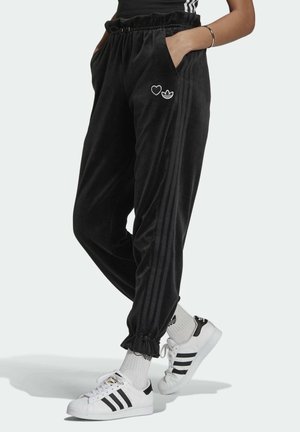 Personne portant un pantalon de survêtement Adidas noir avec un cœur et un logo sur la cuisse, des chaussettes blanches Adidas et des baskets blanches à rayures noires.