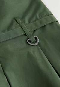Green fabric with a smooth texture features a stitched strap and a metal D-ring. The design includes pleats along the bottom for added detail.