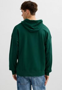 Sudadera verde con un ajuste holgado, puños acanalados y una gran capucha. Se lleva con jeans de mezclilla azul claro. Tela lisa, sin estampados visibles.