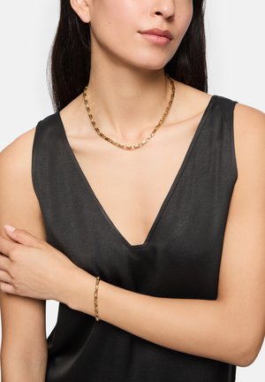 Collier et bracelet en chaîne en or ; maillons rectangulaires ; texture lisse ; portés avec un haut en satin noir à décolleté en V profond.