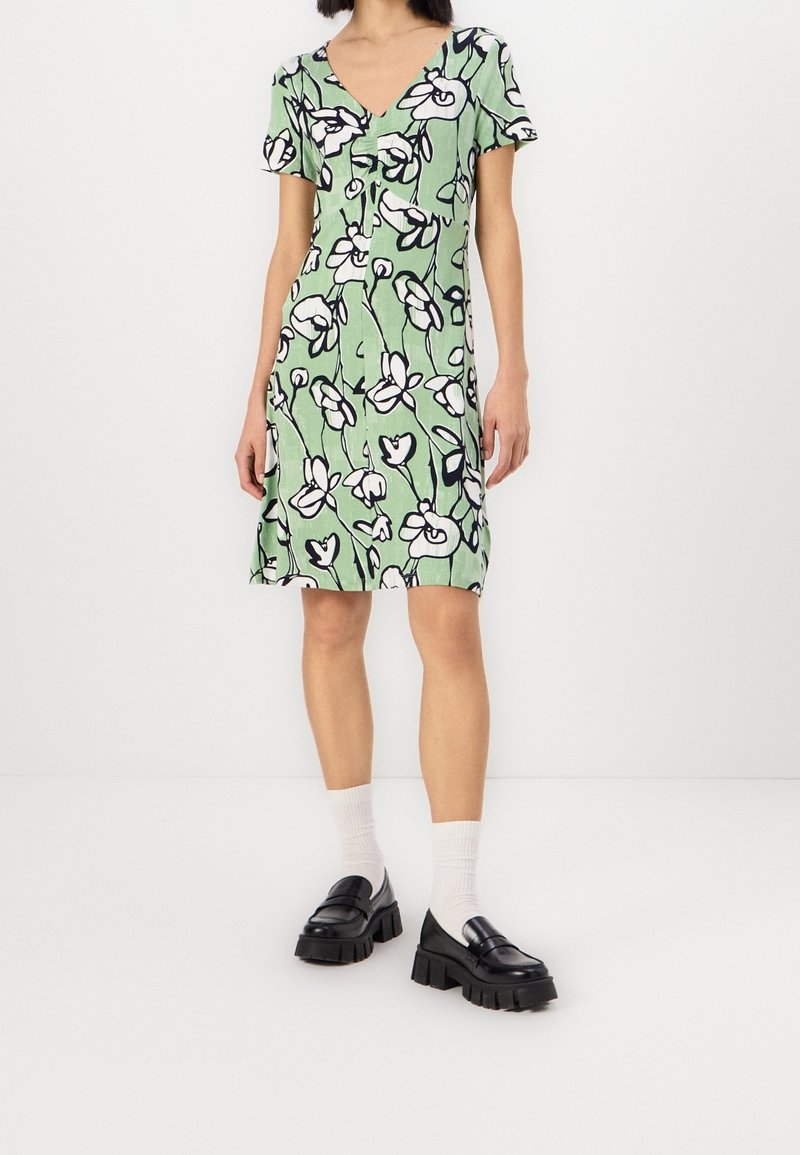 Femme portant une robe verte à manches courtes avec un imprimé floral blanc et noir, associée à des chaussettes blanches et des mocassins noirs épais.