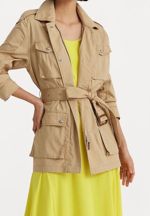 Beige värinen vyöllä varustettu takki, jossa suuret läppätaskut, yllä varattomaton kirkkaan keltainen mekko, malli poseeraa yhdellä kädellä lantiolla.