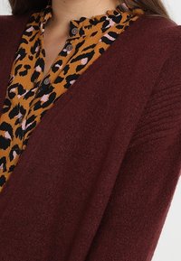 Burgundy stickad cardigan över en gulblommig blus med svart och rosa leopardmönster, som har en volangkrage och knapprad.