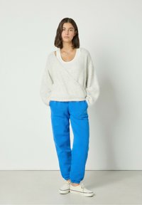Weißer, flauschiger Wickelsweater mit langen Ärmeln, kombiniert mit elektrischen blauen, strukturierten Jogginghosen und weißen Stoffschuhen. Einfacher, geschichteter Look.