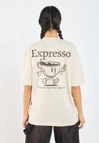 Krémové tričko s krátkým rukávem a grafickým návrhem kresleného šálku kávy, text "Expresso" nahoře a hnědé inkoustové detaily. Uvolněný střih, bavlněný materiál.