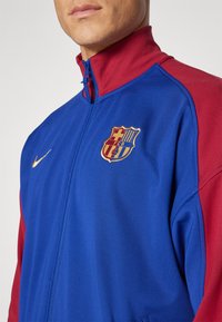 Nike treningsjakke med blått stoff og røde detaljer, høy krage, hel glidelås og brodert FC Barcelona-logo på brystet.