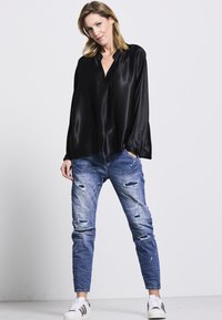 Schwarze Satinbluse mit V-Ausschnitt, kombiniert mit ausgewaschenen blauen Jeans mit Rissen. Weiße Sneaker mit schwarzen Streifen runden das Outfit ab.
