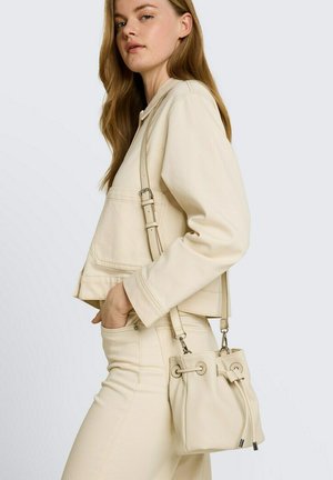 Vrouw in beige jas en broek met een bijpassende bucketbag met trekkoord die schuin over het lichaam wordt gedragen, tegen een effen achtergrond.