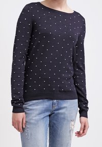 Granatowy sweter w białe kropki, okrągły dekolt i długie rękawy. Dzianinowa faktura z ściągaczami na rękawach i u dołu. W zestawieniu z zniszczonymi dżinsami.