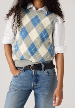 Personne portant une chemise blanche aux manches retroussées, un gilet en tricot à losanges bleu, gris et crème, un jean bleu et une ceinture noire, les mains détendues.