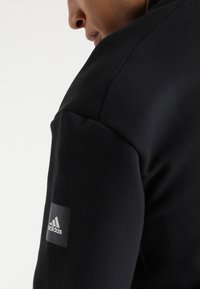 Sweatshirt de sport noir avec une texture lisse, un design ajusté et un patch logo Adidas contrasté sur la manche droite.