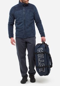 Mężczyzna w granatowej polarowej kurtce, ciemnych spodniach i czarnych butach trzyma granatową torbę sportową z napisem "RILLE".
