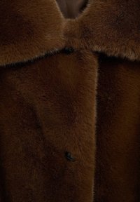 Brązowy płaszcz futrzany o miękkiej, teksturowanej powierzchni, z dużym kołnierzem i zapięciem na jeden guzik. Materiał wydaje się gruby i luksusowy.