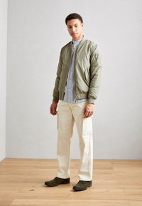 Giacca bomber verde oliva con polsini a costine, camicia azzurra a bottoni, pantaloni cargo bianco sporco e stivaletti verde scuro alla caviglia. Sfondo in legno.