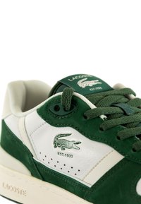 Sneakers Lacoste con una parte superiore in cuoio bianco e dettagli in camoscio verde scuro, chiusura con lacci, marchio logo e suola in gomma testurizzata.