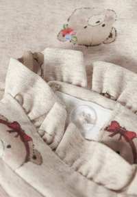 Jasnobeżowy top z materiału, zdobiony uroczym nadrukiem misia oraz kwiatowym akcentem. Falistym kołnierz z zapięciem na guzik z tyłu. Miękka tekstura.