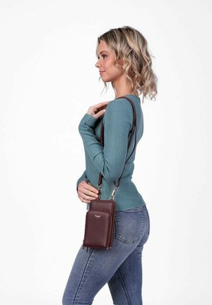 Hnědá kožená taška přes rameno se zipovým uzávěrem a logem, nošená na popruhu přes rameno. Modelka má na sobě zelený žebrovaný top s dlouhým rukávem a džíny.